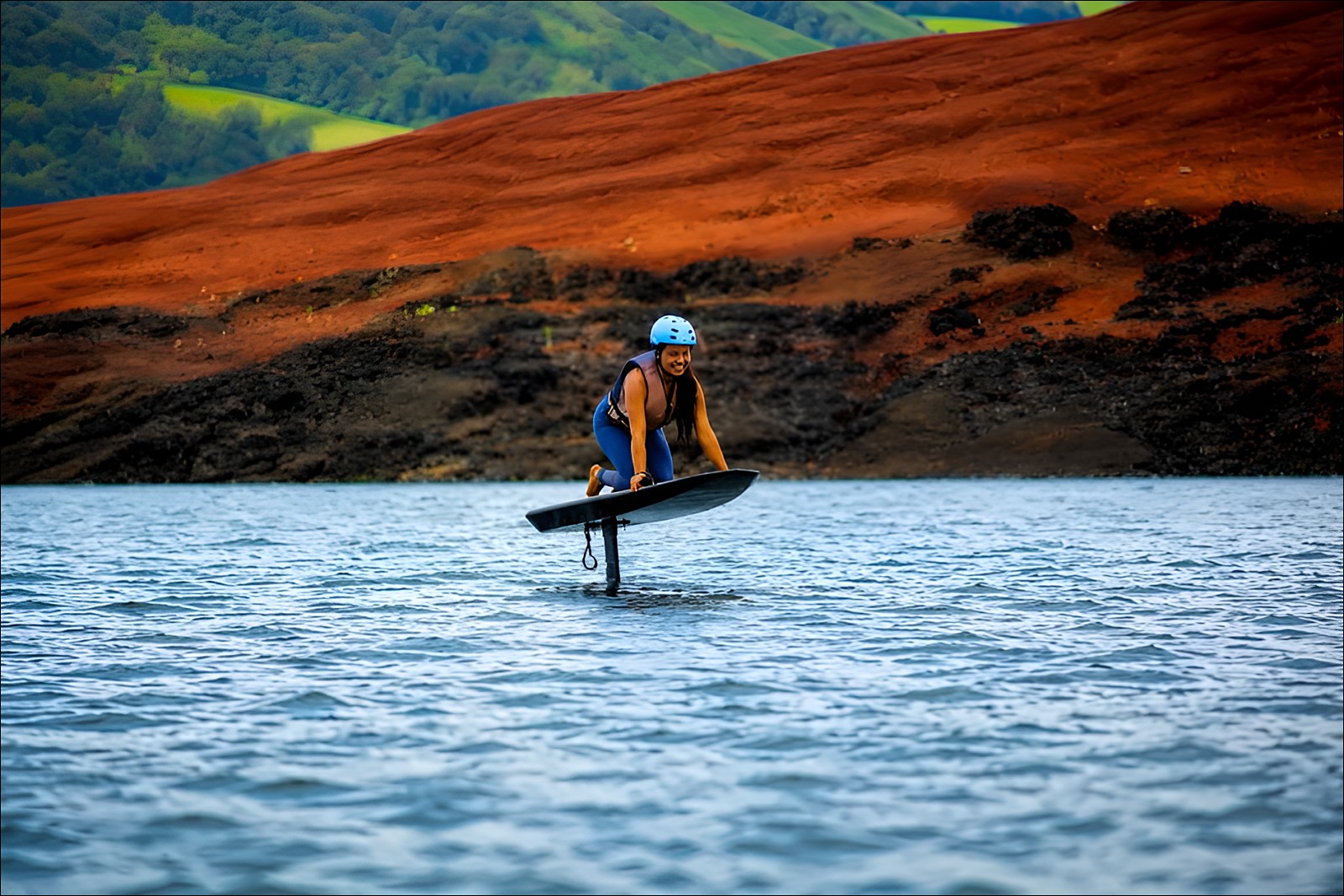Rider principiante con casco y chaleco salvavidas, sonriendo mientras vuela en eFoil sobre el Lago Arenal, con las orillas de tierra roja al fondo